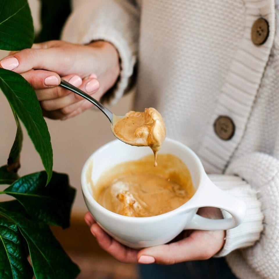 Person in a cream sweater holds a white cup of foamy coffee while lifting a spoonful of creamy foam beside leafy plants.