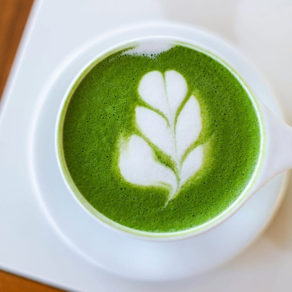 Overhead view of a green matcha latte in a white cup, topped with leaf-shaped foam art on a saucer.