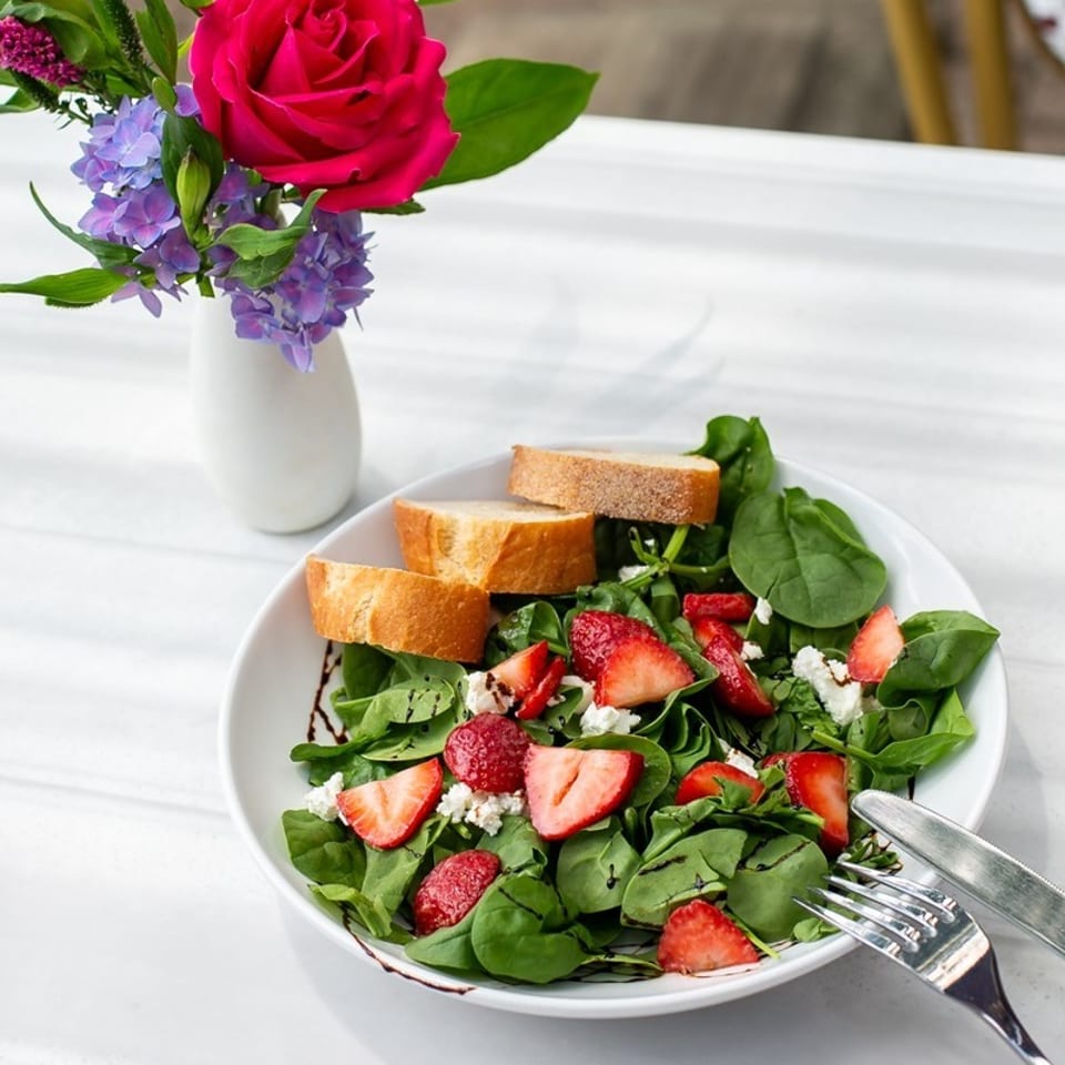 Spinach and strawberry salad with feta, balsamic drizzle, and toasted bread beside a pink rose and purple flower arrangement.