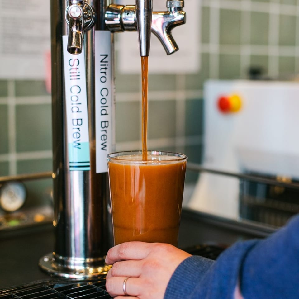 Bartender pours cold brew coffee from a tap into a glass beside labeled still and nitro cold brew taps.