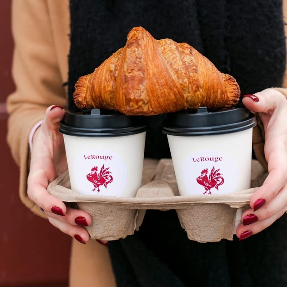 Person holding two LeRouge coffee cups and a golden croissant in a cardboard drink carrier.