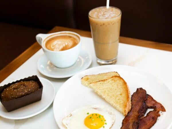 A breakfast spread featuring a fried sunny-side-up egg, crispy bacon strips, toasted sourdough bread, a mini loaf seasonal cake, a cappuccino, and an iced latte at Le Rouge Bakery.