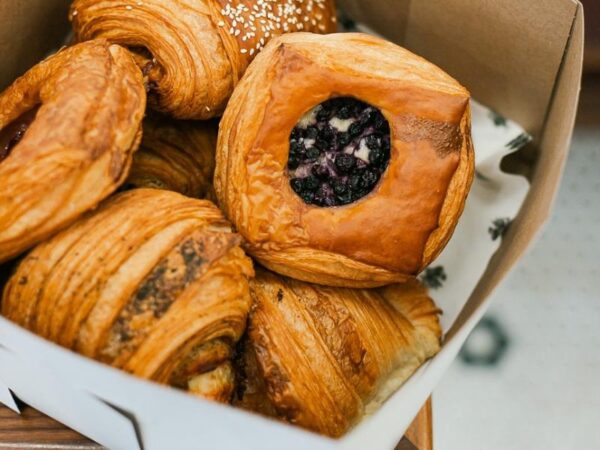 A bakery takeout box filled with fresh flaky pastries, including a round blueberry Danish, seeded pastry, and croissants at Le Rouge Bakery.